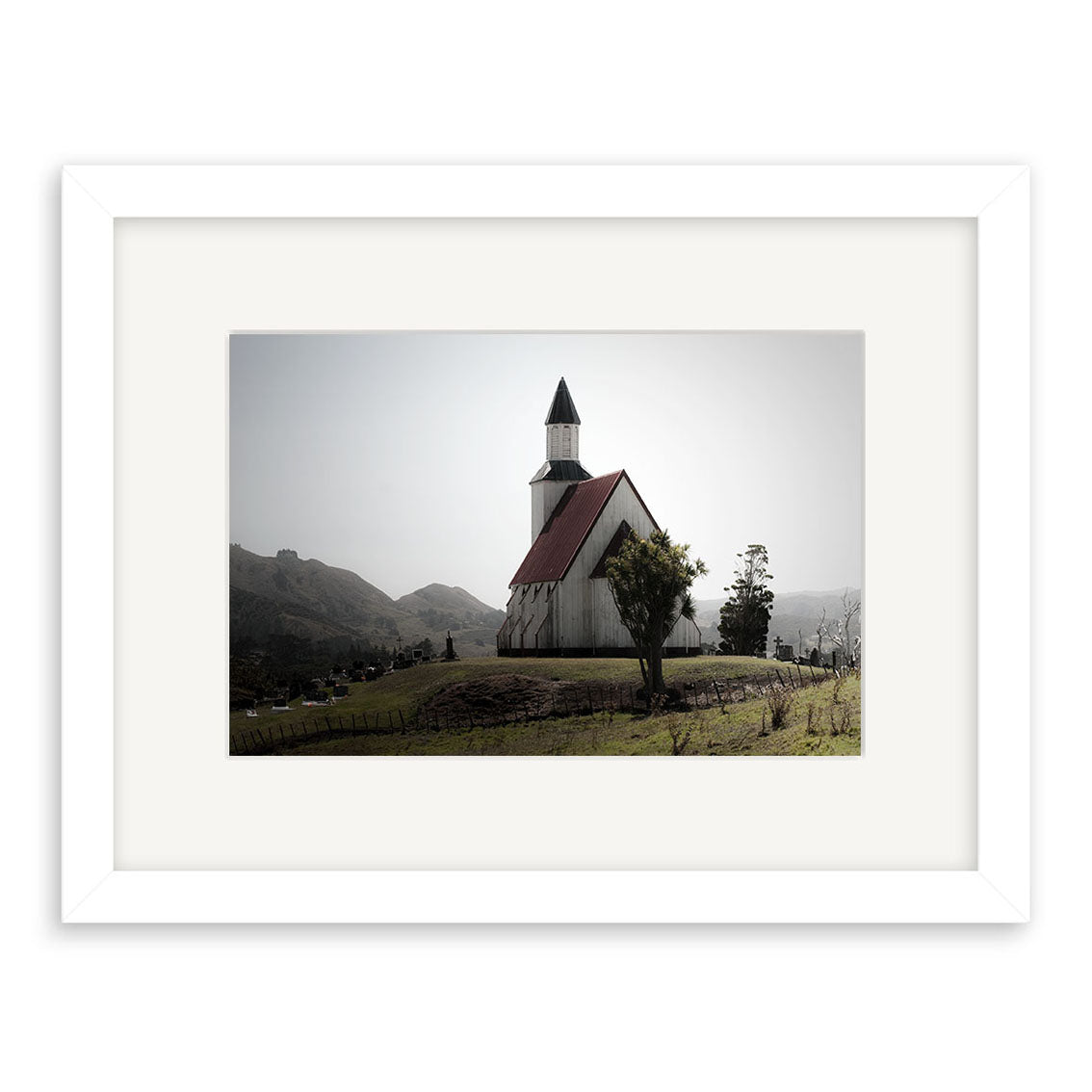 Photograph titled Pawarenga - RP244 of a white church with red roof and steeple on a grassy hill, mountains behind. Captured in New Zealand, available as archival matte prints framed with a white mat and frame.