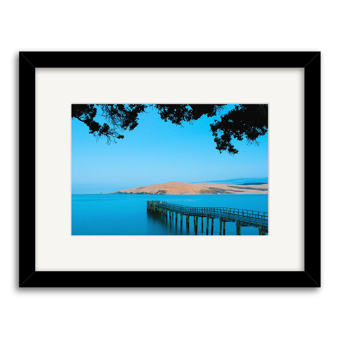 “Omapere – RP245” is a framed photograph by Reuben Price showing a wooden pier reaching over calm blue water toward a distant hill and trees. This New Zealand print is on archival matte paper with a white mat and black frame.