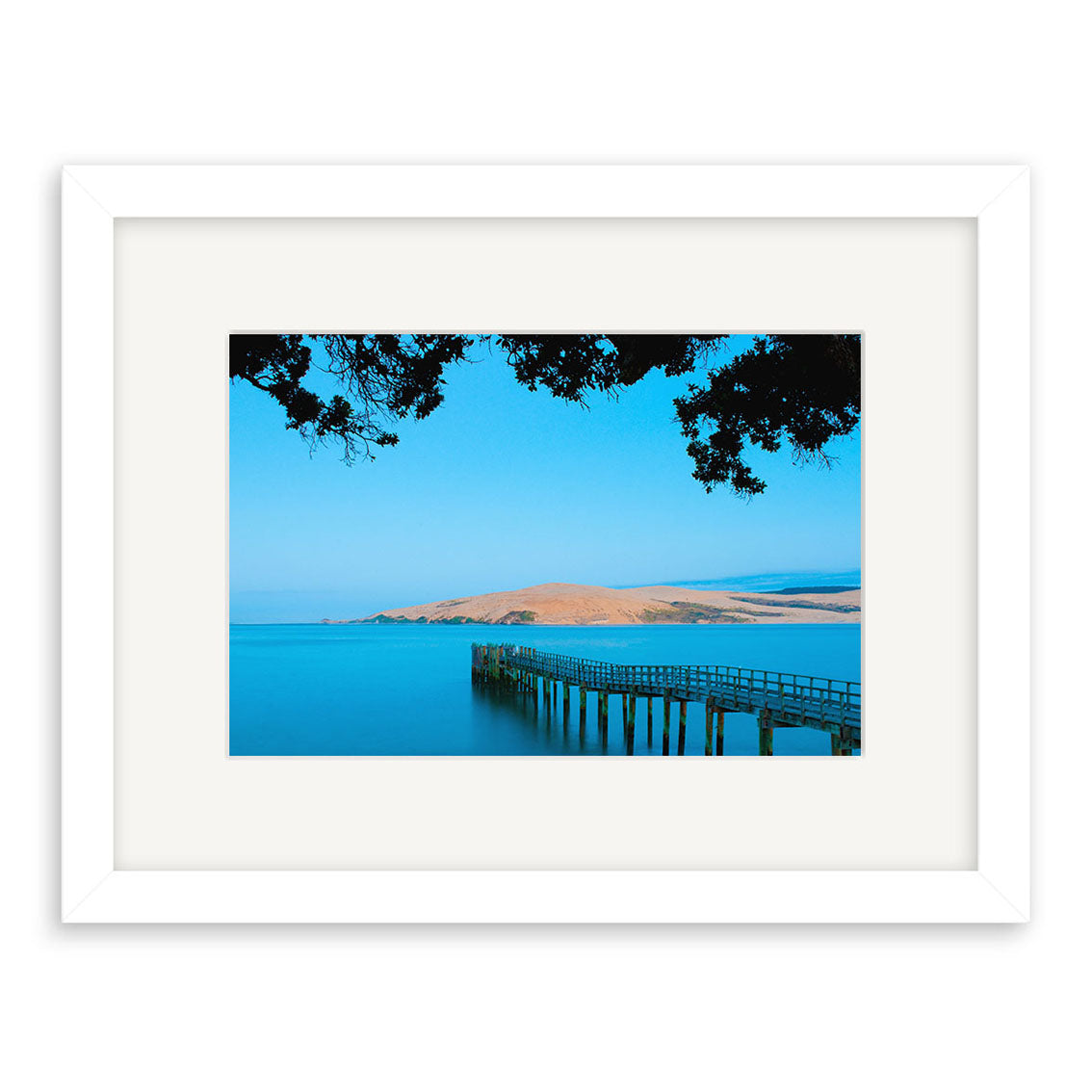 “Omapere - RP245” is a framed photo of a wooden pier reaching into calm blue water, with hilly coastline and tree branches. Part of Reuben Price’s New Zealand collection, it’s printed on archival quality matte paper.