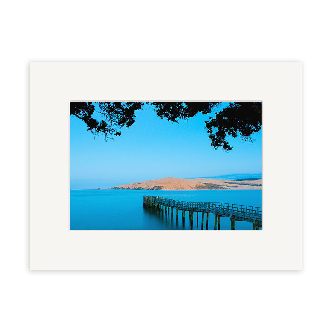 A long wooden pier stretches over calm blue water toward a grassy shore beneath a clear sky, framed by tree branches. Omapere - RP245 by Reuben Price is printed on archival quality matte paper with a wide white mat.
