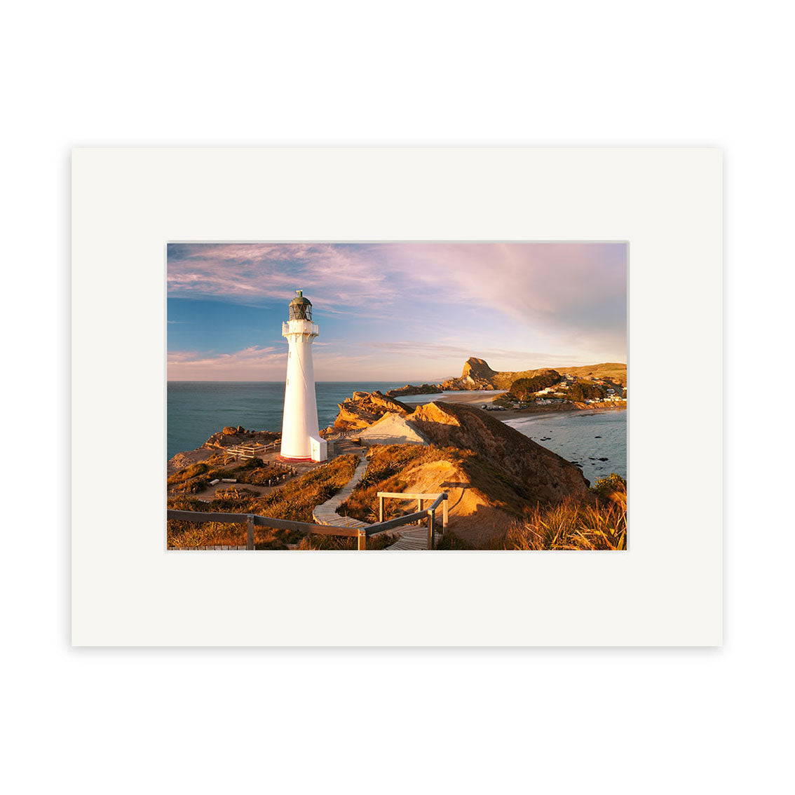 Castlepoint Lighthouse - RP247 stands on a rocky New Zealand coast, overlooking the ocean as a walkway leads to it under soft pink and blue sunset skies—ideal for stunning photographic prints.