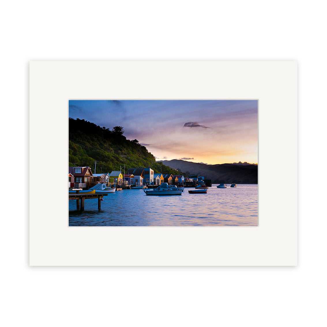 Paremata - RP248 is a photographic print on archival quality paper with a white mat border, depicting colorful houses and boat sheds along the New Zealand waterfront at sunset, with boats and wooded hills in the background.