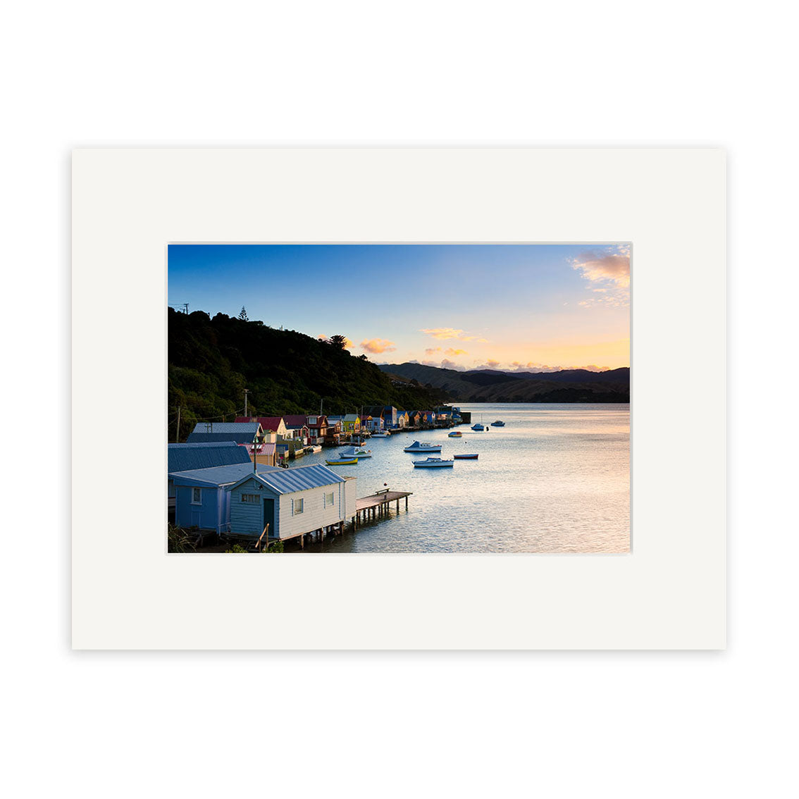 A row of small houses on stilts lines the bay at sunset, bathed in a warm glow—captured in Paremata View - RP249, stunning New Zealand photography by Reuben Price, available as archival quality prints.