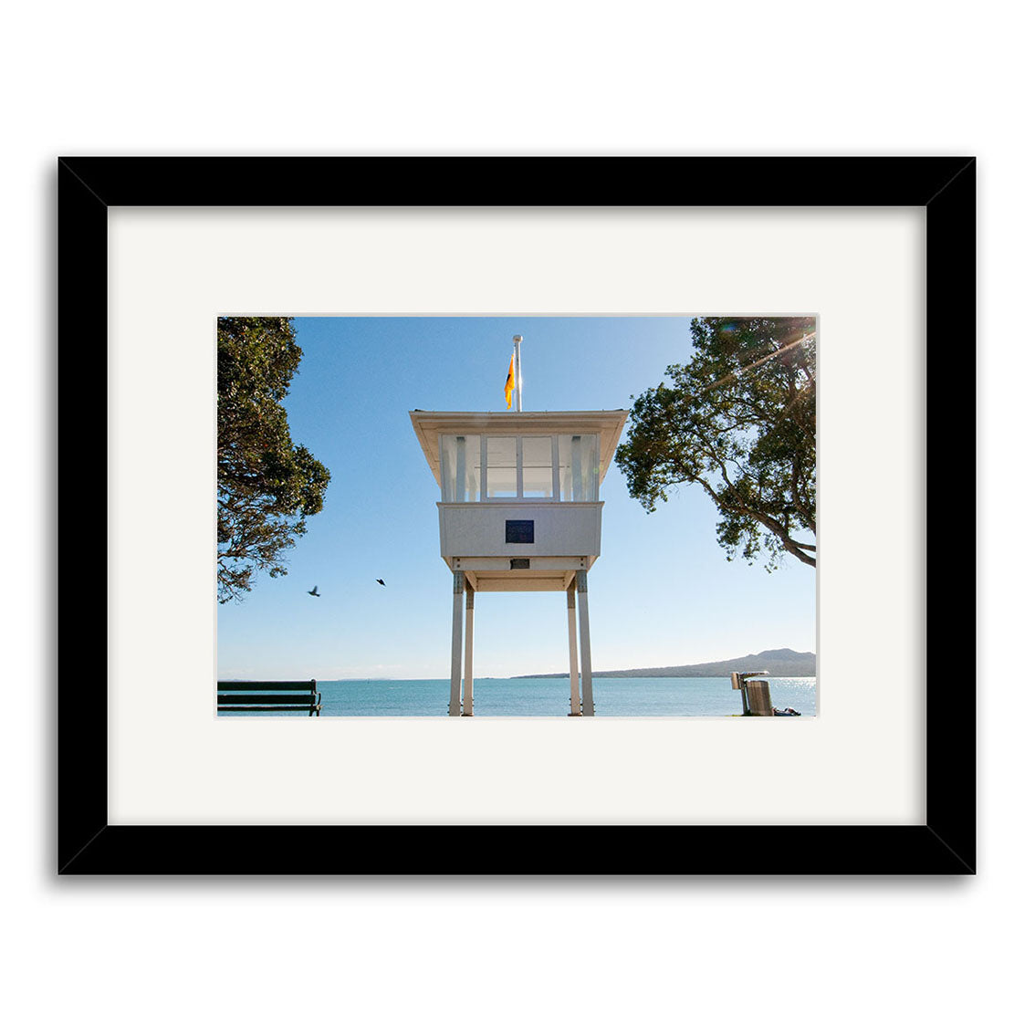 Narrow Neck 2 - RP251 features a framed photo of a white lifeguard tower on stilts by the water in New Zealand, with birds flying nearby, trees on both sides, and a bench—available as archival quality matte paper prints.