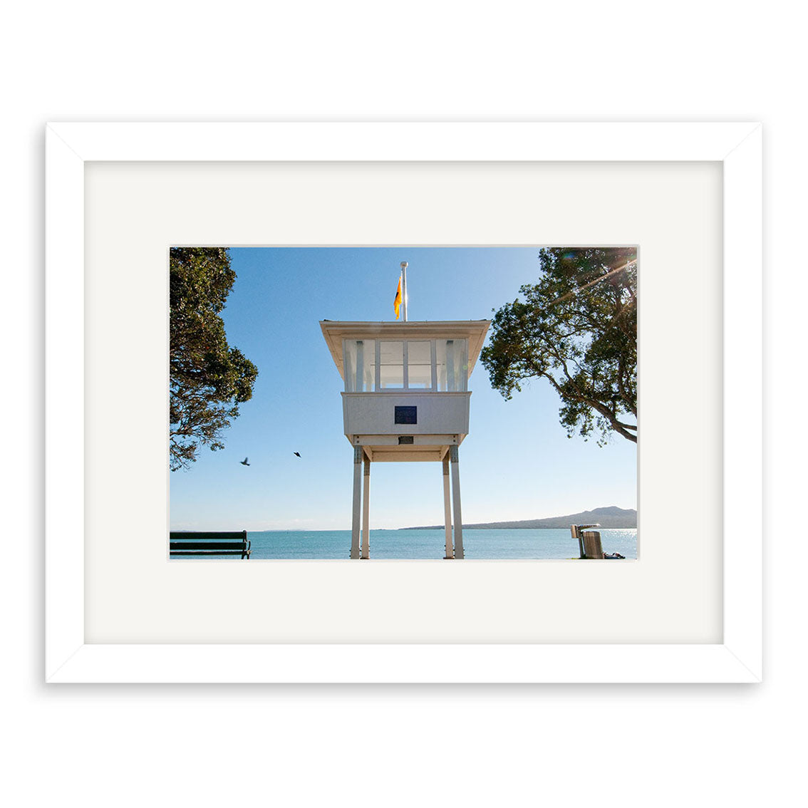 Narrow Neck 2 - RP251 features a framed photo of a white lifeguard tower on stilts by New Zealands water, surrounded by trees and two flying birds under a clear blue sky. Printed on archival quality matte paper for lasting beauty.