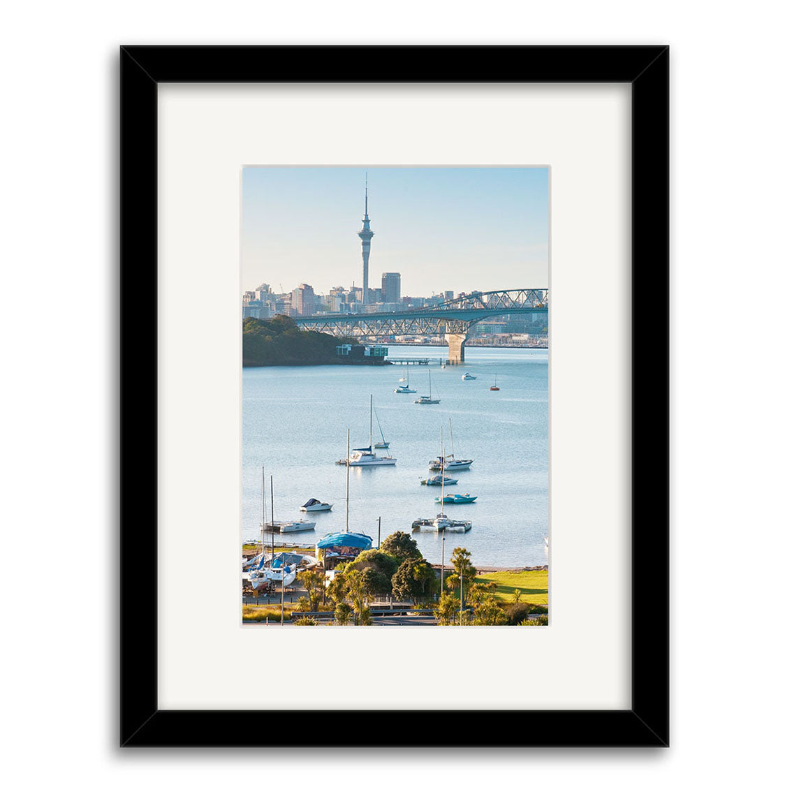 Framed photograph Little Shoal Bay - RP252 features sailboats on the water, a bridge, and city skyline with a tall tower under clear skies. Printed on archival quality paper, this print showcases New Zealand’s coastal charm.