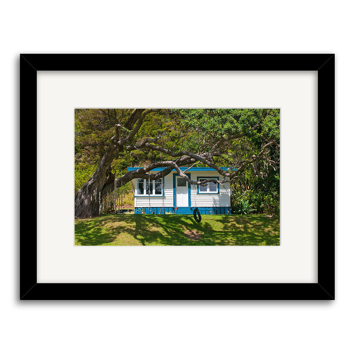 The Helena Bay Bach - RP253 is a photographic print of a small blue and white cottage on a grassy lawn under leafy trees in New Zealand, elegantly framed with a wide white mat and black frame.