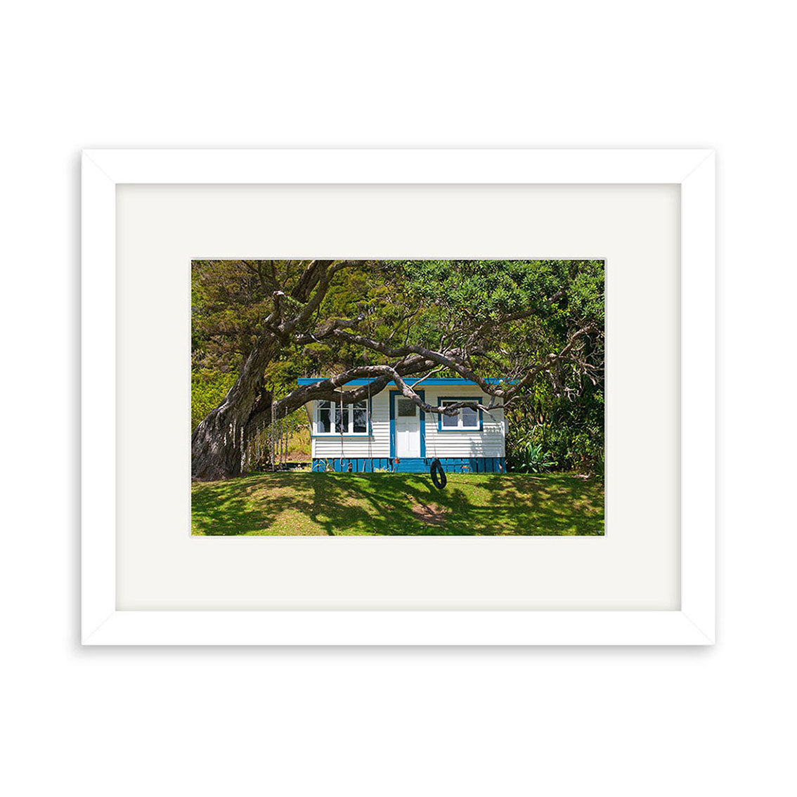 The Helena Bay Bach - RP253 is a framed photographic print of a small white and blue house with a porch, set among green grass and leafy trees. Displayed on white background and printed on archival quality paper for lasting beauty.