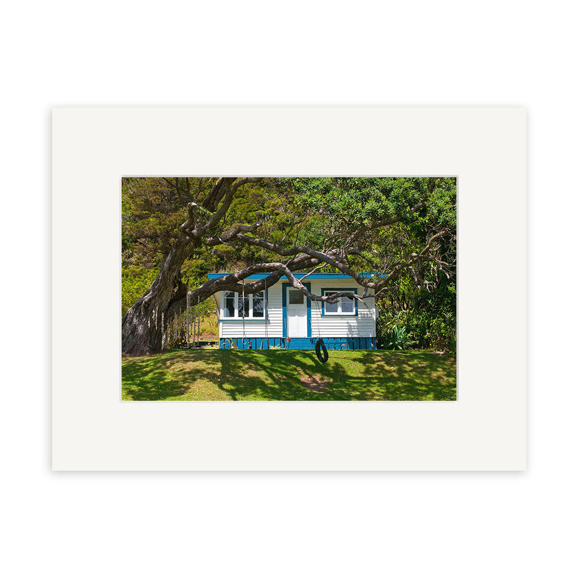 Helena Bay Bach - RP253 shows a small blue and white house with green trim on a grassy lawn beneath leafy trees in New Zealand, available as photographic prints on archival quality paper.