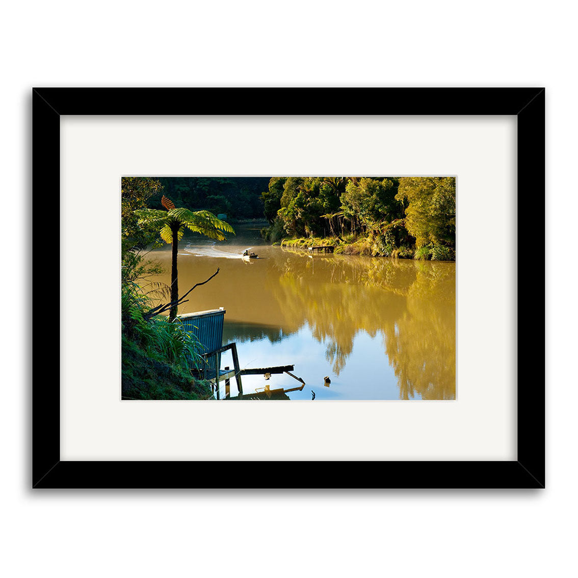 Framed photograph Mokau River - RP257 by Reuben Price, printed on archival matte paper, captures a calm river with green trees and a small boat gliding under soft sunlight near a shed and dock.
