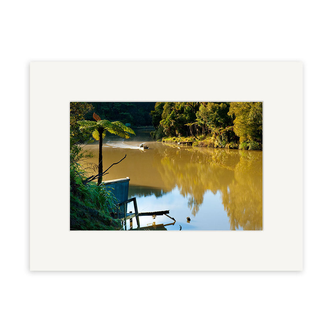 Mokau River - RP257 depicts calm, brownish waters bordered by lush green trees, with a small shed on the bank and a rowboat nearby. This tranquil scene is featured in Reuben Prices renowned New Zealand photographic prints.