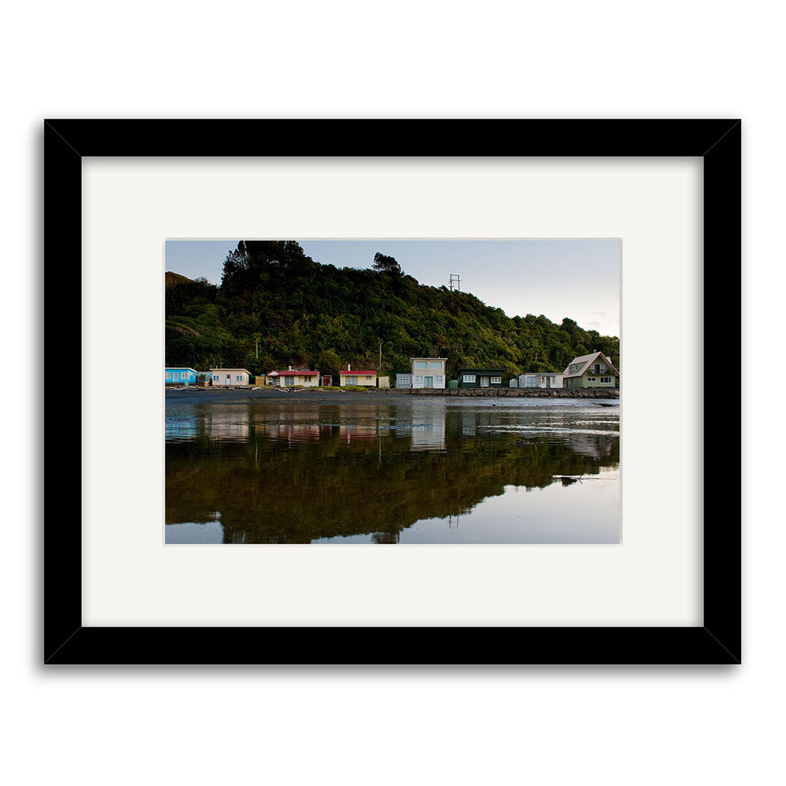 Tongaporutu Baches - RP258: Framed photo of small houses on a New Zealand shoreline with reflections in calm water, hills and trees behind, under a clear sky. Printed on archival quality matte paper for lasting beauty.