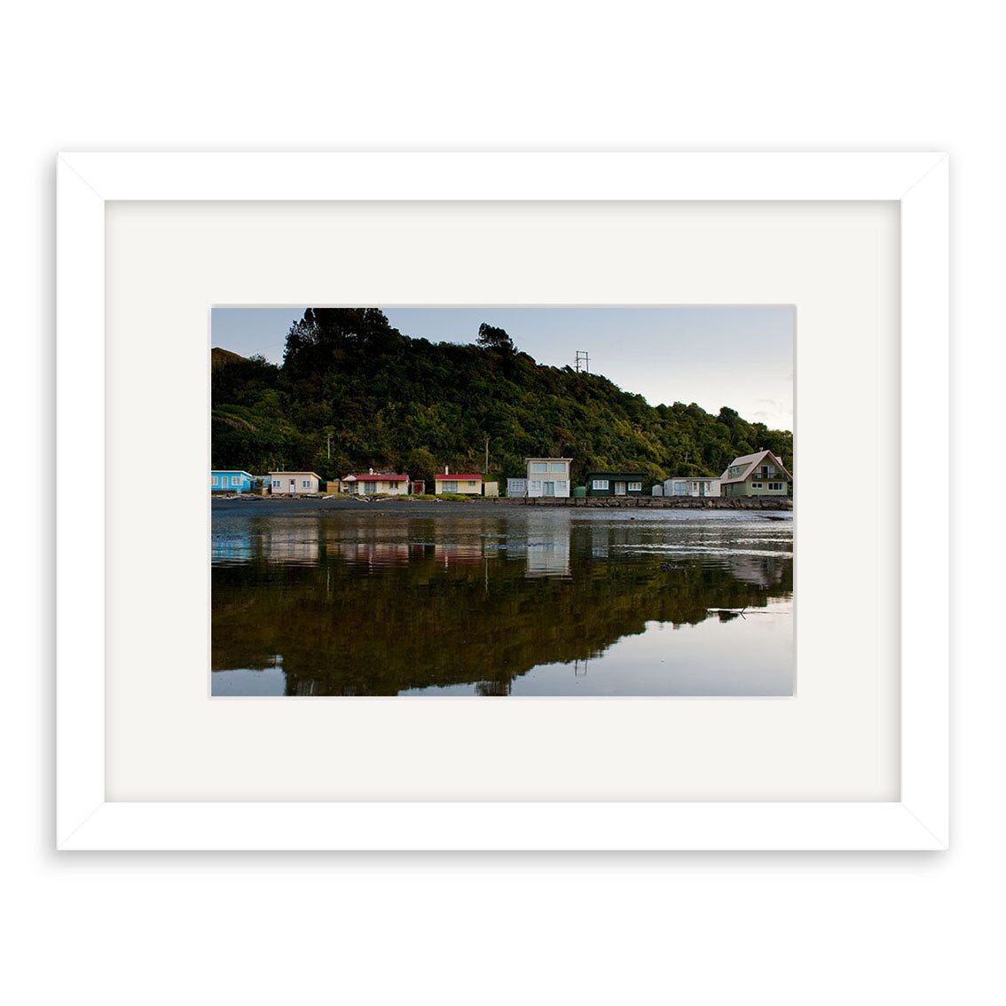 The Tongaporutu Baches - RP258 is a framed photographic print featuring small houses along a New Zealand shoreline with a forested hill behind, their reflections shimmering in calm water. Printed on archival quality matte paper for lasting beauty.
