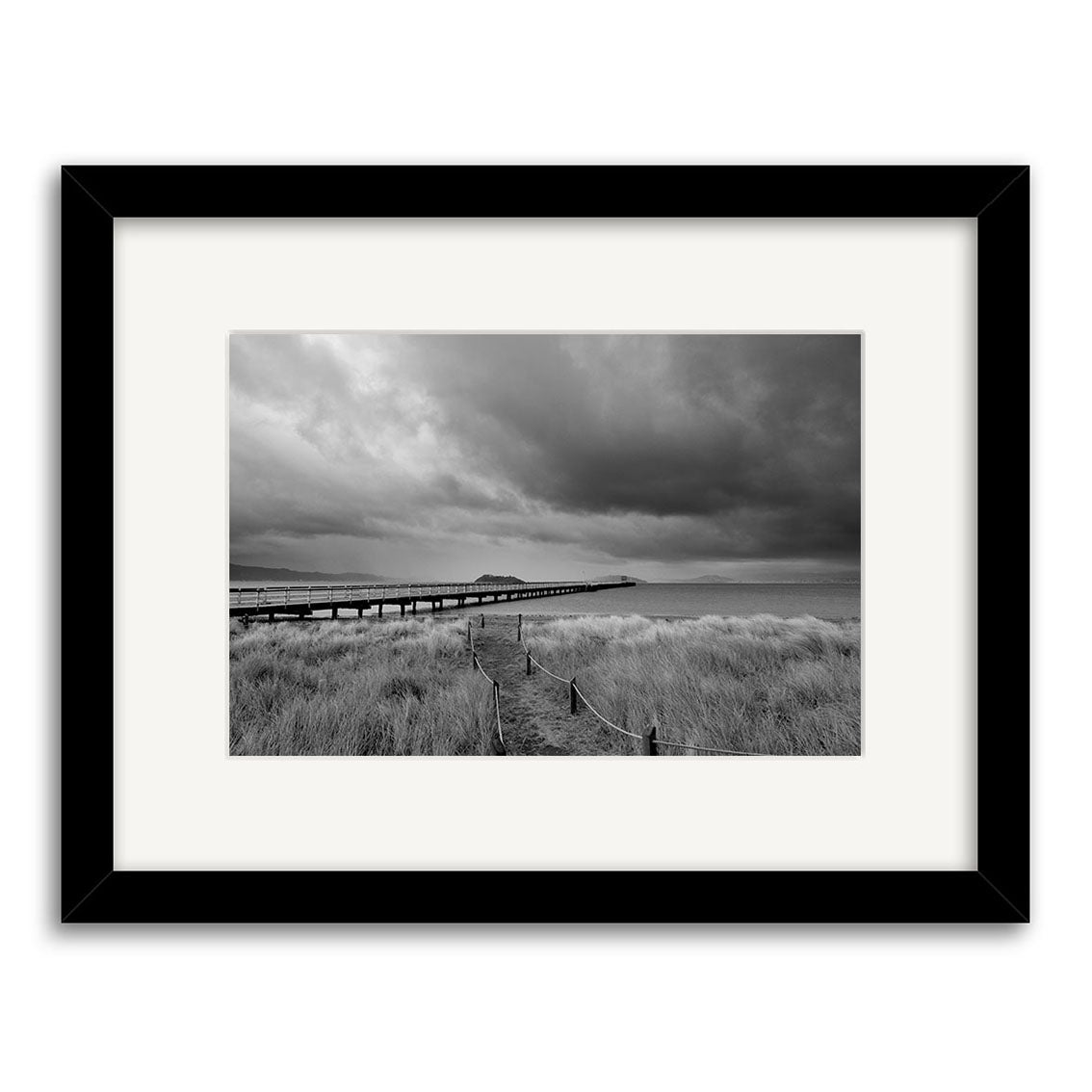 Petone - RP259: Black-and-white photo of a long pier stretching into water, with tall grass in front and a cloudy sky above. Framed in black with a white mat, available as archival quality matte photographic prints.