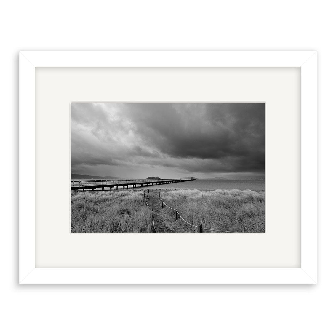 Petone - RP259: Black and white photo of a wooden boardwalk over water in New Zealand, with tall grass and dramatic clouds. Beautifully framed in white mat and frame, printed on archival quality matte paper.