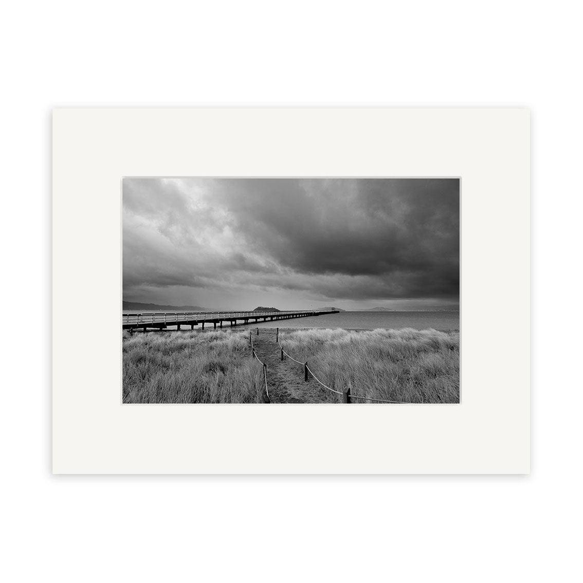 Petone - RP259: Black and white photo of a long wooden pier in New Zealand, with tall grass and cloudy skies. This tranquil scene is available as archival quality matte paper photographic print.