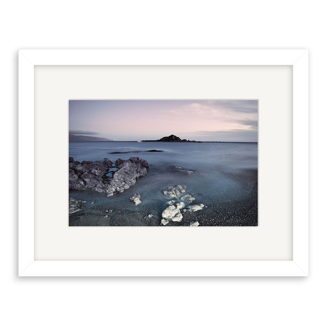 Island Bay - RP260 is a framed archival print featuring a tranquil New Zealand seascape with rocky foreground, smooth water, and a distant island beneath a muted, cloudy sky.