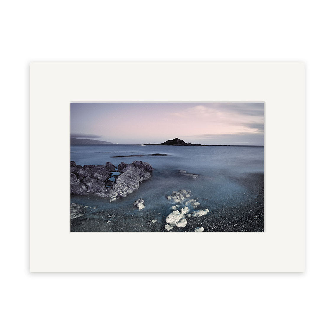 Island Bay - RP260 captures a serene New Zealand coast at dusk with rocks, smooth water, and a distant island under a cloudy sky. This archival quality photographic print comes elegantly framed with a wide white mat.