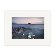 Island Bay - RP260 captures a serene New Zealand coast at dusk with rocks, smooth water, and a distant island under a cloudy sky. This archival quality photographic print comes elegantly framed with a wide white mat.