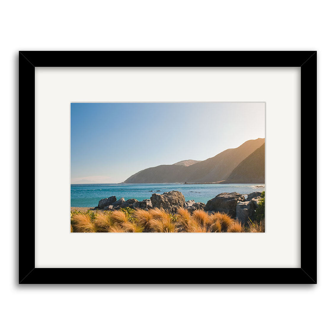 Owhiro Bay - RP261: A framed photograph of a New Zealand coastal landscape with blue ocean, rocky shore, grassy foreground, and distant mountains under a clear sky, printed on archival quality paper.