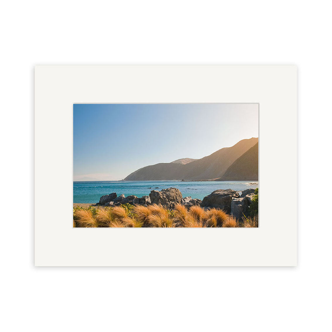 Owhiro Bay - RP261 is a matted photo print on archival paper featuring a coastal landscape with rocky shore, tall grasses, blue ocean, distant mountains, and a clear New Zealand sky.