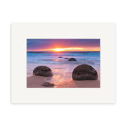 Moeraki Boulders - RP262: A vivid sunset over a New Zealand beach, waves lapping at the sand and large round boulders beneath an orange, pink, and purple sky—ideal for archival quality photographic prints.