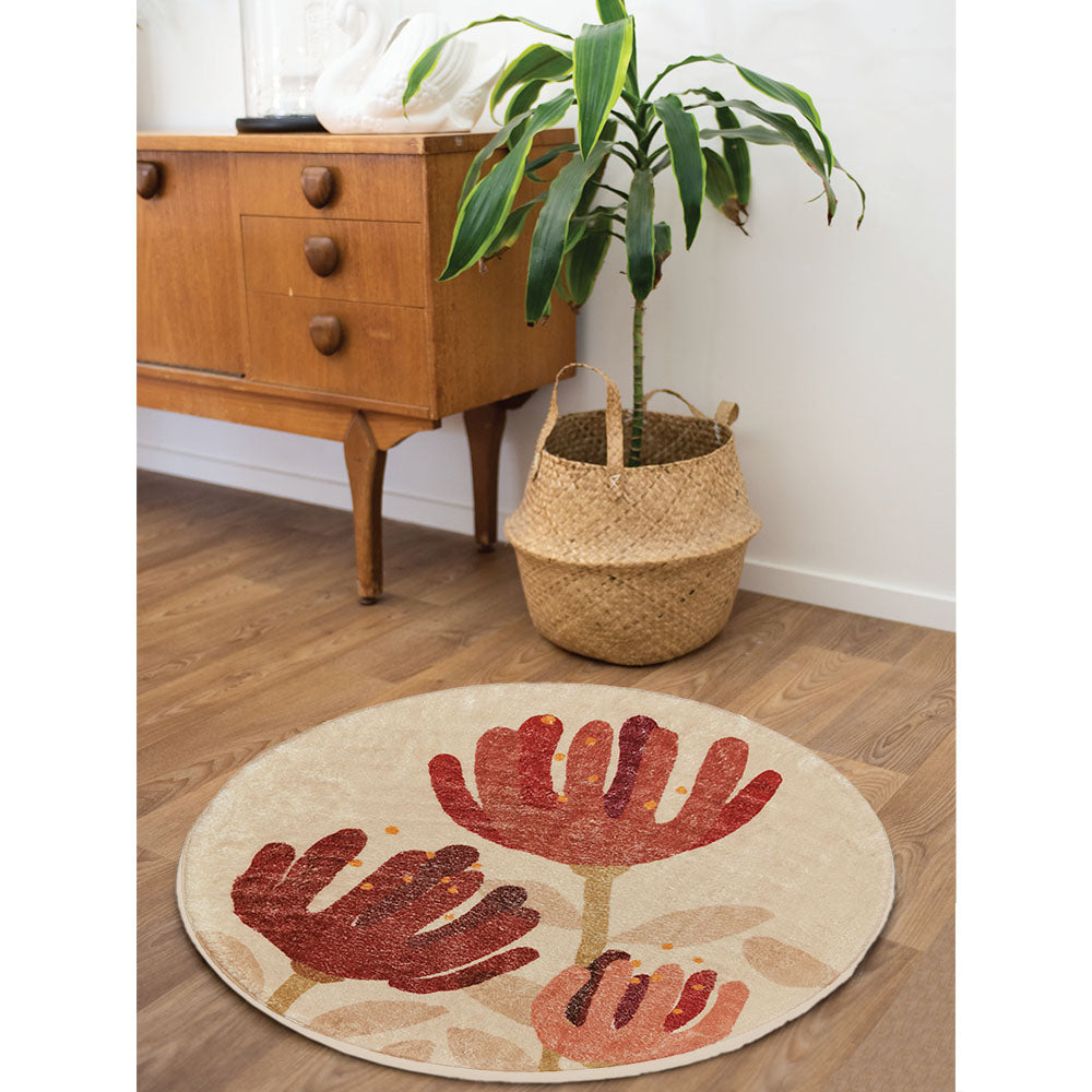 The Raw Pohutukawa Rug, a super-soft round floor rug with red flower designs and anti-slip backing, rests on a wood floor near a mid-century cabinet. Nearby, a tall green plant in a woven basket sits next to the rug.