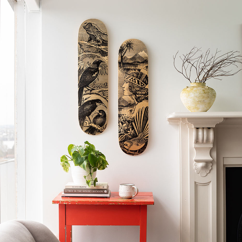 Two Wahine Stamp Skateboard Decks, made from Canadian maple and featuring black-and-white nature illustrations, hang above a red table with a potted plant and candle. A yellow vase with twigs sits on the white mantelpiece to the right.