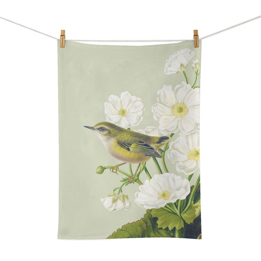 The Birds & Botanicals Rifleman Tea Towel features a small yellow bird among white flowers and green leaves on a pale background, celebrating New Zealand nature. It hangs on a white rope with clothespins for display.