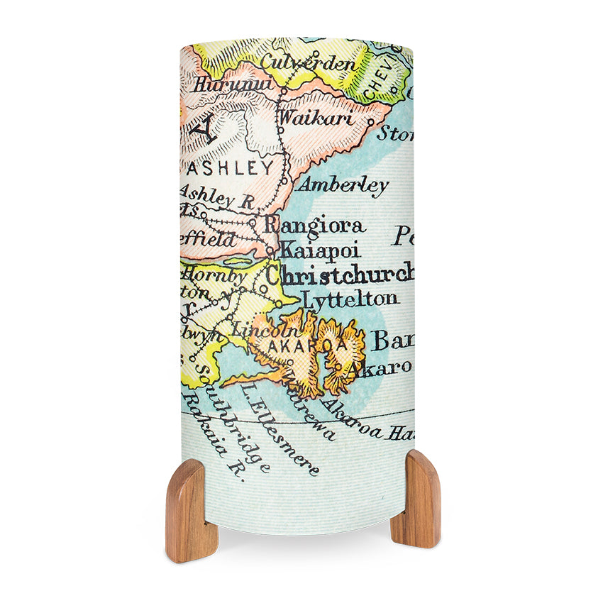 The Vintage Canterbury Map Table Lamp features a cylindrical design with wooden legs and displays a vibrant printed map of New Zealand’s Canterbury region, highlighting cities like Christchurch, Akaroa, Rangiora, and nearby areas.
