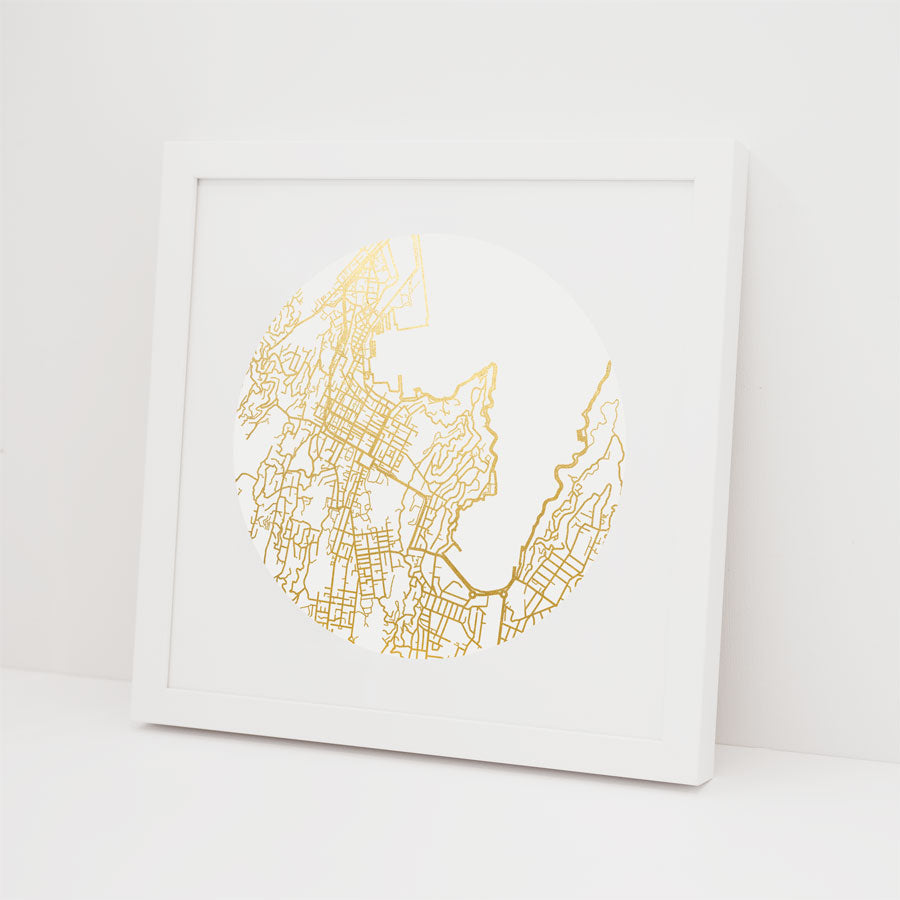 The Wellington Mapscape Foil Print is a framed artwork featuring a circular map design with gold foil street lines on a white background, shown leaning against a white wall.