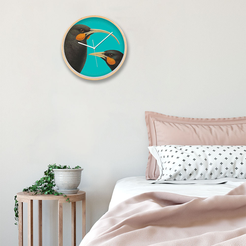 A modern bedroom displays the Bright Huia Wooden Frame Clock with silent movement and a bold huia design, artwork of two black birds on teal, a wooden side table holding a potted plant, and a neatly made bed with pink and white bedding.