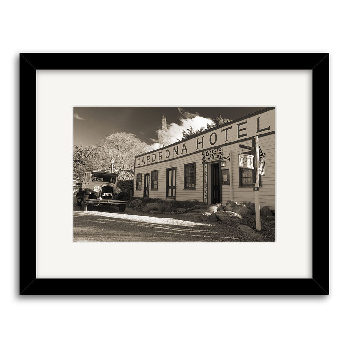Cardrona Pub - RP071: A black-and-white framed photo of New Zealand’s Cardrona Hotel, with an old car and the Cardrona Hotel sign out front. Features trees, clouds in the background. Printed on archival quality matte paper.