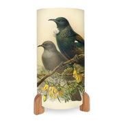 The Buller Tui Table Lamp is a cylindrical lamp with wooden feet, showcasing vibrant artwork of two birds on a flowering branch—one with iridescent green feathers and a white throat tuft—and bright yellow flowers below.