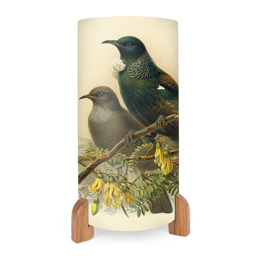 The Buller Tui Table Lamp is a cylindrical lamp with wooden feet, showcasing vibrant artwork of two birds on a flowering branch—one with iridescent green feathers and a white throat tuft—and bright yellow flowers below.
