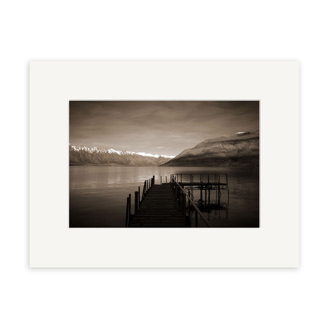 Lake Wakatipu Wharf - RP066: A sepia photo of a wooden pier stretching into a tranquil New Zealand lake, mountains and clouds behind. This print is matted with a white border on archival quality matte paper.