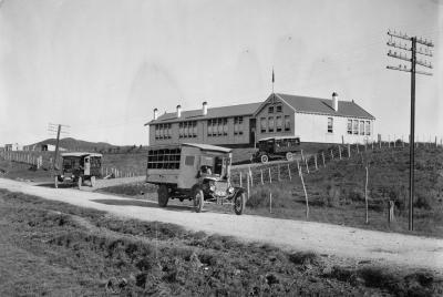 The archival-quality Piopio School Waitomo 1924 No.2 - HI192 print shows three early 20th-century vehicles on a dirt road by a fenced, single-story school atop a hill, with utility poles, open land, and distant New Zealand hills in view.