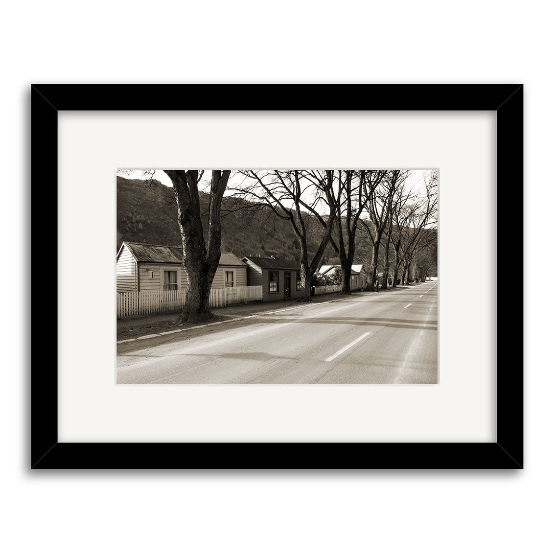 Arrowtown - RP068 is a framed black and white photographic print depicting a tranquil New Zealand street lined with leafless trees and mountains beyond. Archival quality ensures this serene scene remains beautiful for years to come.