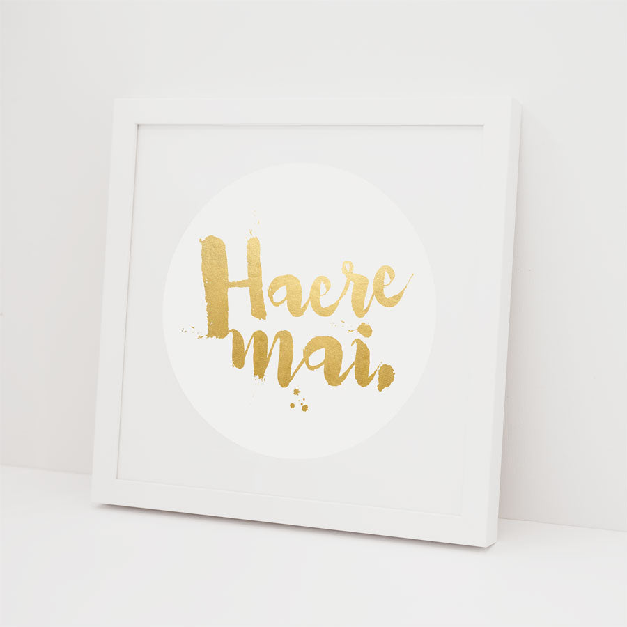 The Haere Mai - Foil Print features gold text on a white background with a white frame, leaning stylishly against a white wall—a modern Kiwiana accent for your home.