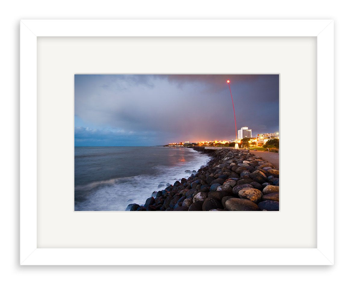 The New Plymouth - RP241 is a framed photograph of a rocky New Zealand shoreline at dusk, with waves, city lights, and a red light trail in the cloudy sky—printed on archival matte paper.