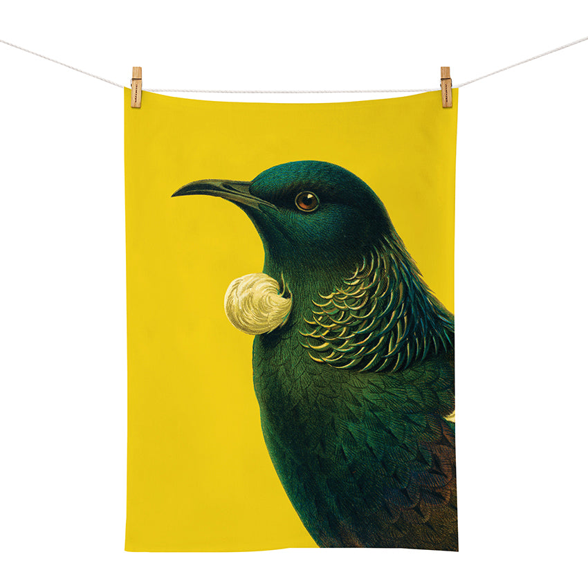 The Bright Tui Tea Towel, screen printed with a tūīs dark iridescent feathers and white throat tufts, hangs from a clothesline against a plain yellow background.