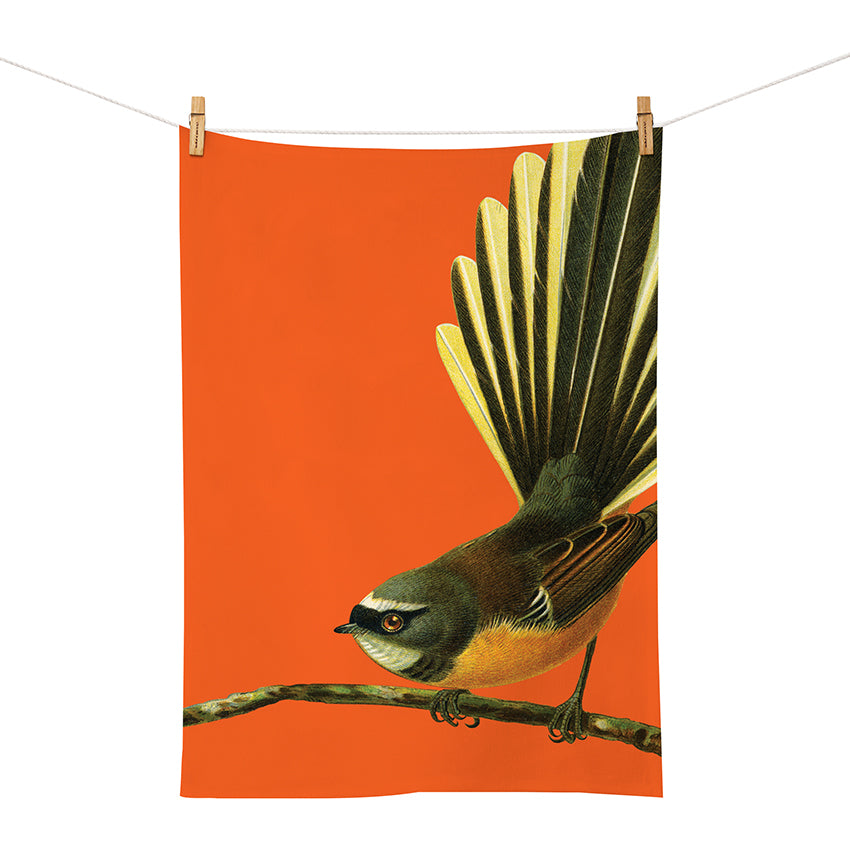 The Bright Fantail Tea Towel is a screen printed cotton towel from the Bright Birds Range, featuring a small bird with fanned tail feathers on an orange background, displayed on a clothesline with wooden clothespins.