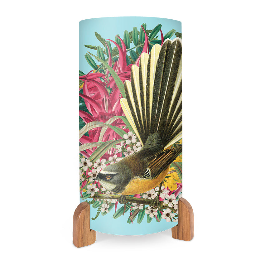 The Botanic Fantail Table Lamp features a cylindrical shape with a wooden base, showcasing vibrant images of a fantail bird amid pink flowers and green leaves on a light blue background—perfect for bringing New Zealand-inspired charm to any room.