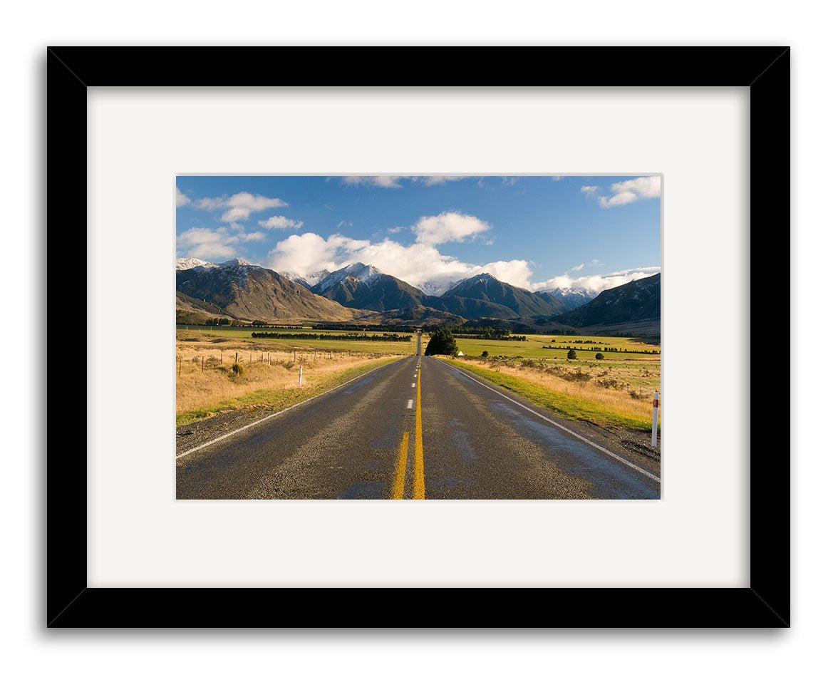 A straight, two-lane road with yellow lines leads into the distance beneath a blue sky. Fields and mountains frame the scene. This black and white photographic print, Arthurs Pass - RP177, evokes New Zealands open roads.