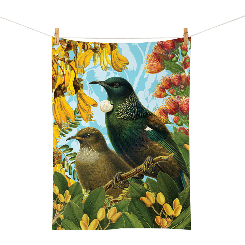 The Botanical Tui Tea Towel features two iridescent green and white birds perched amid red and yellow flowers, printed on cotton. Clipped to a clothesline against a blue sky, its perfect for adding botanical charm to your kitchen.