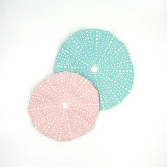 The Kina Bowl Covers (Set of 2) feature pink and blue pastel sea urchin-inspired designs with white dots and central holes, resembling delicate cotton texture, shown overlapping on a white background.