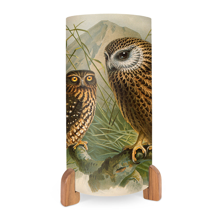 The Buller Ruru Table Lamp is a cylindrical lamp with vivid images of two brown owls on a branch, green leaves, and mountains. It stands on wooden feet and includes an in-line switch at the base for easy use.