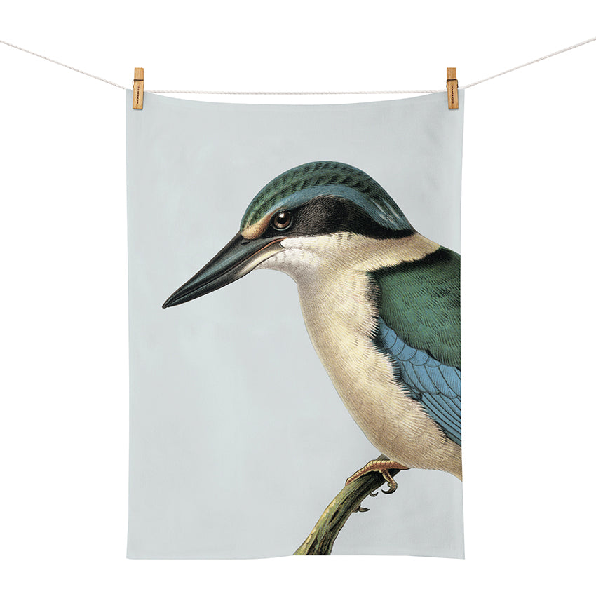 The Hushed Blue Kingfisher Tea Towel displays a printed kingfisher on a branch, hanging from two wooden clothespins. Designed in New Zealand by Hushed Range, it stands out against a simple background.