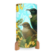 The Botanical Tui Table Lamp has a cylindrical shape, wooden base, and displays vibrant Tui birds among green leaves and yellow flowers on a light blue background, drawing inspiration from New Zealand’s natural beauty.