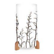 The Harakeke Table Lamp is a cylindrical white lamp with black tree branches and birds, featuring an in-line switch and two wooden base feet.